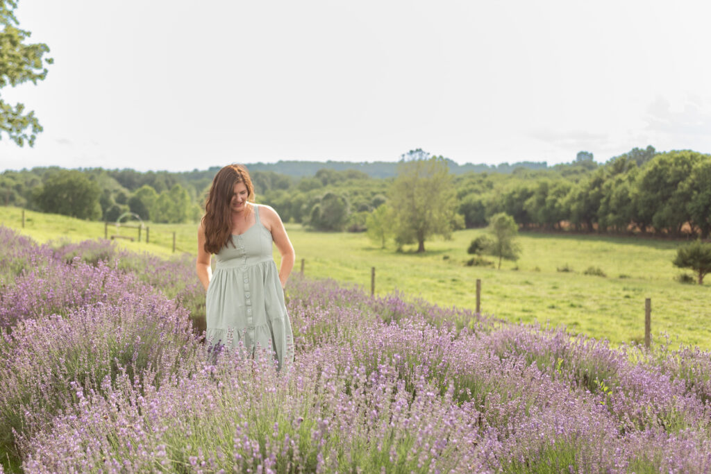 All about my sessions! Photographer guiding clients during a calm, outdoor photo session; Wendy walking through a field of lavender on a bright summer day. She is a brunette, wearing a foam green summer dress with her hands in her pockets, looking down at the ground. 